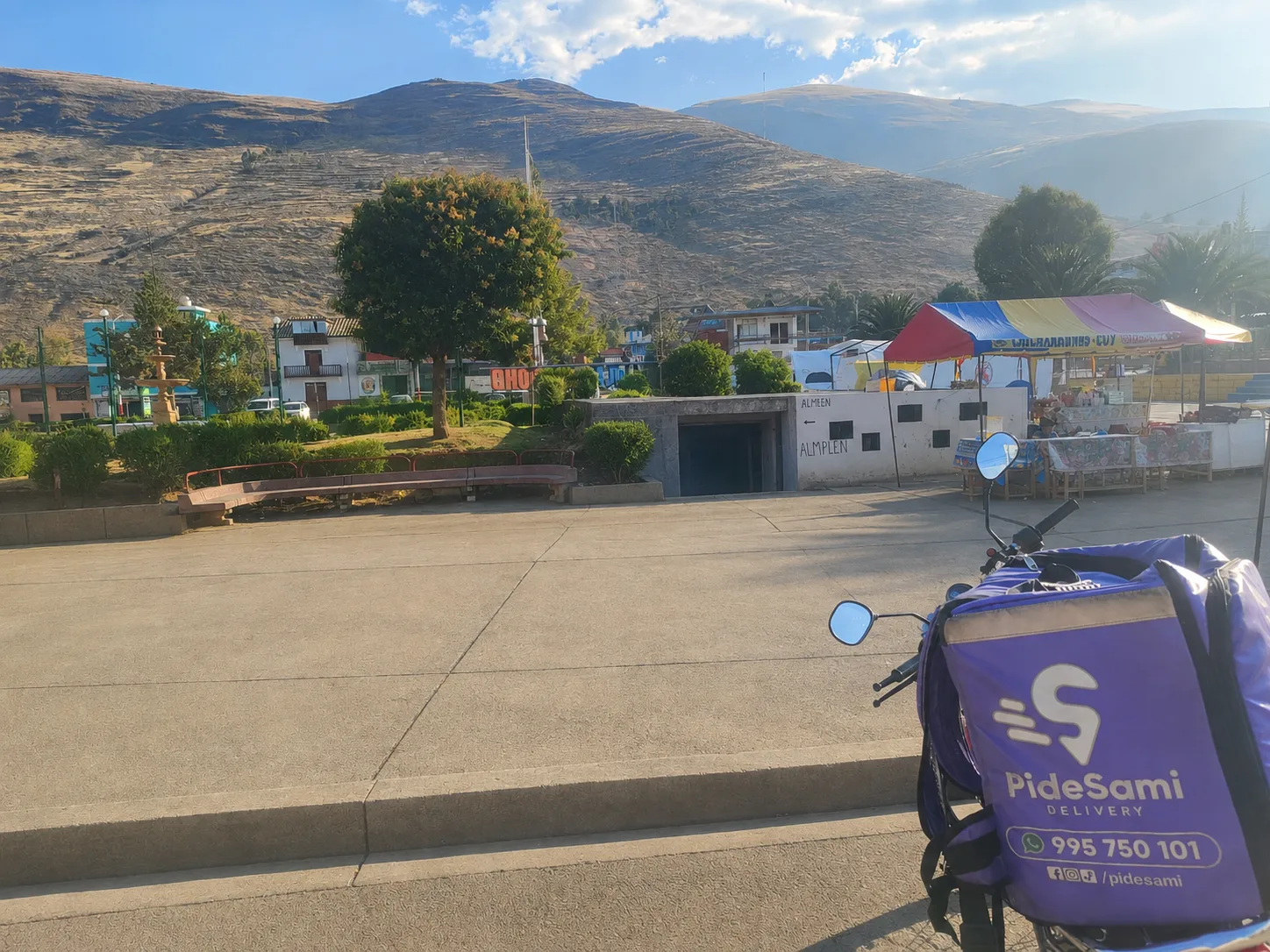 Mochila morada de Pidesami sobre una motocicleta frente a una plaza y el paisaje andino